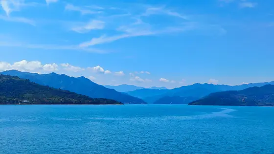 澄澈的蓝铺满天际，几缕游云如仙子遗落的白纱，轻拂过山的眉梢。湖水凝作一块温润的蓝玉，风过时漾起细碎的银鳞，将远山的倒影揉成淡墨。层叠的青山像列队的巨人，披着翠绿的衣袍，守着这一汪碧波的清梦。岸边的绿意探进水里，仿佛要与蓝绸共舞，连呼吸都染上了清润的风。