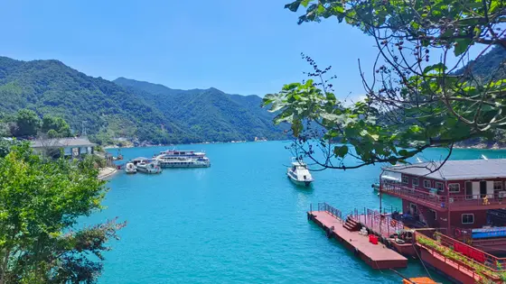 青山如黛环抱着一湖碧玉，阳光把湖面染成流动的翡翠。几艘游船静静泊在水面，白墙黛瓦的屋舍沿岸而生，像是从山水长卷里走出来的渔村。枝叶间漏下细碎的光斑，风掠过时，连倒影都在轻轻摇晃。