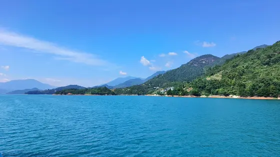 湖水如蓝宝石般闪耀，波纹轻诉着夏日的低语。青山环抱，绿意盎然，几间小屋静静依偎在岸畔。远山在薄雾中若隐若现，天际线与云朵共舞。整片天地静谧而悠远，仿佛时间在此刻凝固。