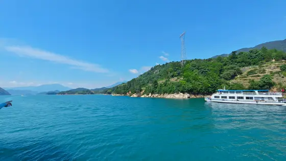 碧空澄澈，几缕闲云悠然舒展，似天空轻落的羽。湖水蓝得透亮，微风拂过，碎银似的鳞光在水面轻颤。岸边青山叠翠，绿意浓稠得像要溢出，与碧波温柔相依。白船轻摇，载着一湖诗意，正滑向那片被阳光吻过的山湾。远处山峦蒙着薄青纱，唯有高塔静立，成了天地间最沉稳的注脚。