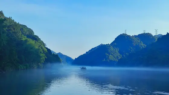 轻纱般的晨雾漫过江心，将远山晕染成一抹青黛。一叶扁舟悄然划过，仿佛载着时光的呼吸。两岸青山如屏，倒映在澄澈的水面，与天光云影共徘徊。