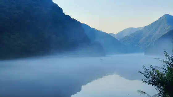 清晨的薄雾如轻纱般覆盖在静谧的江面上，远山青黛如眉，隐约可见。水面倒映着山影，虚实交织，仿佛一幅流动的水墨画。一叶扁舟在雾中缓缓行进，船上人影静默，似与天地融为一体。天色渐明，微光穿透云层，洒在粼粼波光之上，宁静中透出无限遐想。