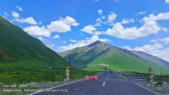 柏油路蜿蜒伸向远方，像一条墨色丝带系在青山之间。头顶是澄澈如洗的蓝天，白云朵朵如棉絮般悠然飘浮。远处的山峦披着翠绿的绒毯，在阳光下泛着温柔的光泽。风过处，仿佛能听见云朵与山风的低语。这里，天地相接处，便是人间最美的远方。