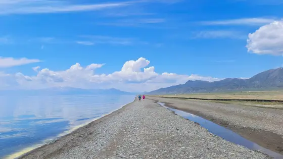 天空的蓝倒映在赛里木湖的眸中，云朵轻舒成岸边远山的絮语。两个人沿着碎石小径漫步，仿佛走进一幅被晨光浸透的水彩画。湖水以永恒的沉默收纳天光，每一片波纹都写着高原宁静的诗篇。