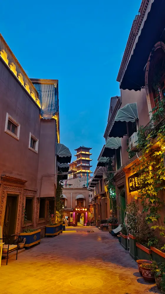 暮色轻垂，巷陌间灯火温柔地苏醒。砖瓦的纹理在暖光里低语，像被时光抚过的旧信笺。远处塔影静立，仿佛一盏点亮苍穹的灯，将整条街巷拥入怀中。