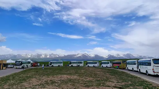 蓝天下，云朵如轻纱般舒卷，远处雪山连绵，静静矗立在天际线上。碧绿的草原铺展开来，一列大巴缓缓停驻，像是为这片宁静添上了一笔人间的暖色。风轻轻吹过，带着山野的气息，让每一次驻足都成为与自然的对话。