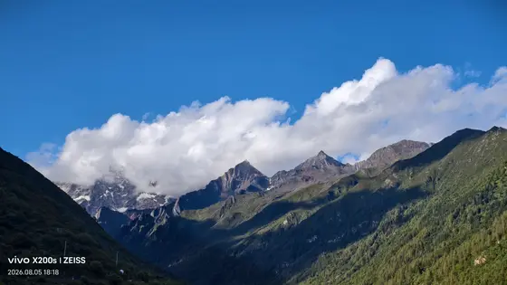 天光倾泻在蜿蜒的山脊上,云雾如轻纱半掩着雪峰的眉眼。风在深谷间游走,将松涛的私语藏进墨绿的皱褶里。那片积雪的山巅仿佛沉睡的巨人,正缓缓掀开云被,露出亘古的苍茫。山脚下的森林却悄悄苏醒,用无数翠绿的掌心,接住从峰顶滑落的流光。