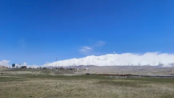 湛蓝如洗的天幕下，绵延的雪山披着银装，宛如沉睡的巨龙。山脚下的草地轻铺绿毯，稀疏的脚印指向远方。游人拾级而上，红色的旗帜在风中轻扬，仿佛与天地共呼吸。这片静谧的辽阔，将喧嚣隔绝于云端之外。