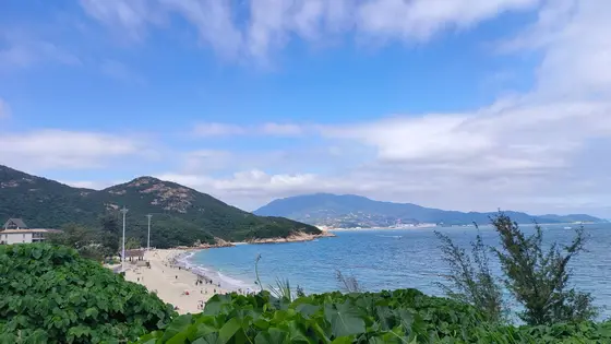 天光云影徘徊在蔚蓝的海面上，青黛的山峦温柔地拥抱着一弯银白的沙滩。海风轻拂过婆娑的树影，将远山的轮廓勾勒成一幅淡墨画卷。沙滩上散落的笑语，仿佛是时光里轻轻洒下的音符，与涛声和鸣成一曲自然的韵律。
