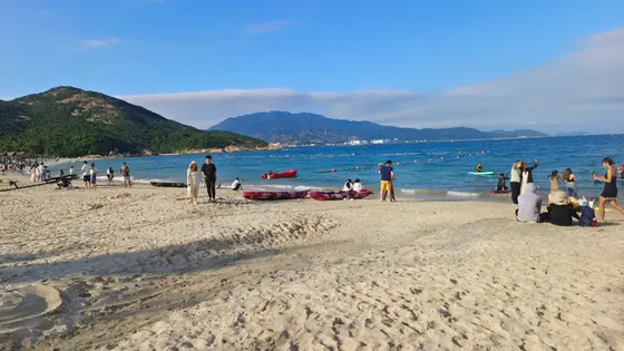 潮水轻抚沙岸，留下蜿蜒的诗行。碧波之上，人影与小舟点缀着时光的涟漪。远山如黛，在云絮间舒展，守护着这片蔚蓝的宁静。