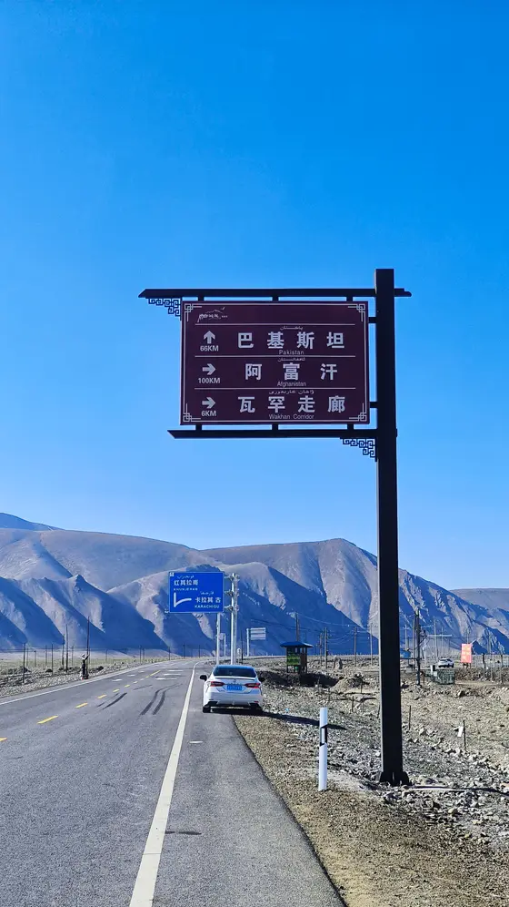 苍穹如洗，蓝色的天幕下，路牌静默地指向远方——巴基斯坦、阿富汗、瓦罕走廊，几个名字沉甸甸地压在风里。笔直的公路切开莽莽山峦，一辆白车正驶向那片辽远的未知，仿佛旅途本身就成了诗行。