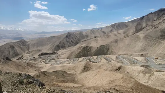 苍茫山脊如大地的皱褶，一条银蛇般的公路在褐色峡谷间迂回攀升。云影掠过荒芜的山坡，远方雪峰静卧在天际线。孤独的车辆如音符般点缀在蜿蜒的旋律里，丈量着高原沉默的厚度。