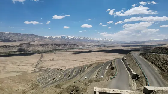 公路如一条沉睡的巨龙，蜿蜒着伸向雪山的王座。  
云影在荒原上流淌，几朵闲云是天空写下的诗行。  
车流是时光的碎屑，沿着山的脊梁，缓缓滑向天际。  
此刻，世界只剩下蓝与褐的对话，静得能听见风的叹息。