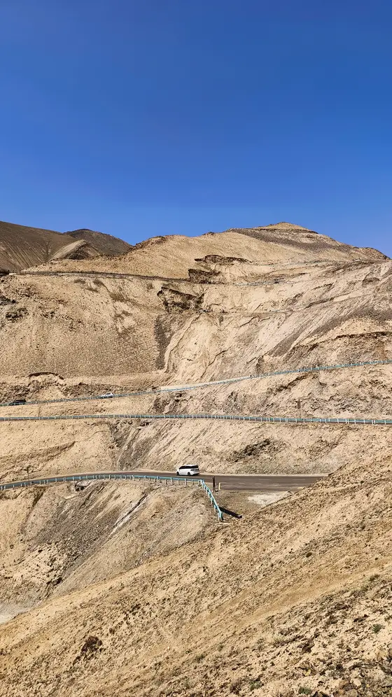 群山如凝固的浪涛，在高原静默起伏。  
公路是大地掌心蜿蜒的掌纹，托起一抹流动的云白。  
车影渺小如一粒尘埃，驶向天际线那一抹幽蓝的缝隙。  
风沙磨亮了岩壁的褶皱，时间在此处缓慢呼吸。