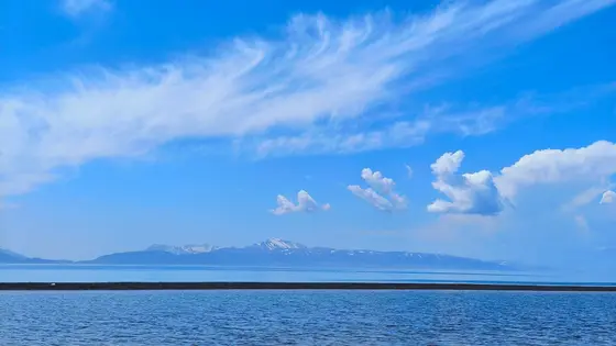天空如一块澄澈的蓝水晶，几缕流云似轻纱般舒展，有的如羽扇轻摇，有的如浪卷云堆。远处的雪山披着薄雪，在蓝天下静默伫立，似与天边的云絮低语。湖水蓝得深邃，与天相接处泛着柔波，一道暗色的岸线横卧水中，像大地轻触水面的指尖。风很轻，云很慢，时间在这里慢成了永恒的蓝。