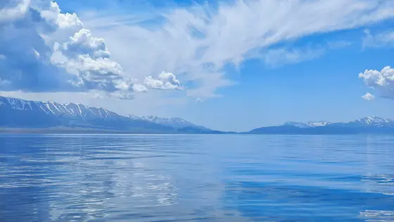 湖水如镜，倒映着雪山的轮廓，仿佛天空洒落的颜料在水面晕染。云朵悠然舒展，似轻纱拂过蔚蓝的穹顶，又似游龙在湖心留下斑驳的影。远山静默，却以雪线为笔，在天地间勾勒出永恒的韵律。