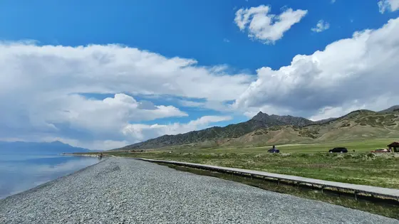 天空铺展着淡蓝的绸缎，云朵轻轻停泊，像写给山峦的短笺。湖水收容了天光与峰影，碎石滩低语着潮汐的往昔。木栈道蜿蜒向远处，仿佛在邀请风，去阅读青草与山脊的私语。