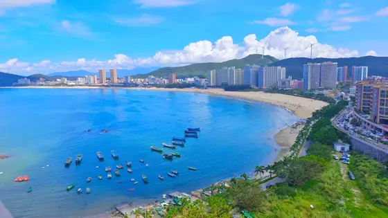 晨光为海面铺上蓝绸，渔船如散落的棋子静卧波间。云朵在远山之巅堆叠成雪，高楼与椰影相映，勾勒出海与城温柔的相遇。风从风车叶隙间漏下细语，将沙滩上零星的欢笑揉碎在潮声里。