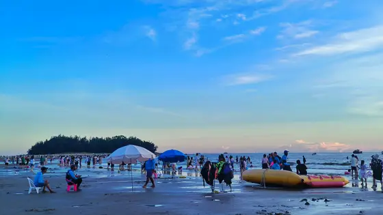 暮色温柔地铺展，将天空染成渐变的绸缎，云朵如絮，轻缀着蔚蓝与浅金的边界。沙滩上散落着欢声笑语，脚印与浪花私语，伞下的剪影拉长了时光的温柔。那静卧的充气船，仿佛是海风遗落的梦，等待下一次潮起时，载着星辉漂向远方。