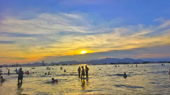 暮色如轻纱垂落，海面铺展着碎金的涟漪。  
夕照将云絮染成暖黄，远山在薄雾中晕成一抹淡蓝。  
嬉水的人影剪成诗行，欢声融进波光，与归鸟共赴夜的序章。