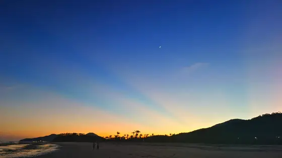 暮色如一块渐变的绸缎，从天顶的靛蓝滑落到海面的橘黄。一弯细月悬在空阔处，像谁遗落的银簪。远山与椰林的剪影沉默着，将沙滩上两枚细小的影子，温柔地衬入这片将暗未暗的天地间。